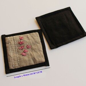 Puede incluir: Dos posavasos cuadrados de tela, uno con la parte superior de tela negra y otro con la parte superior de tela beige con flores bordadas de color rosa. El posavasos beige tiene un borde negro y el texto "Longitud x Ancho: 12CM*12CM".