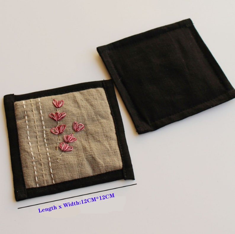 Puede incluir: Dos posavasos cuadrados de tela, uno con la parte superior de tela negra y otro con la parte superior de tela beige con flores bordadas de color rosa. El posavasos beige tiene un borde negro y el texto "Longitud x Ancho: 12CM*12CM".