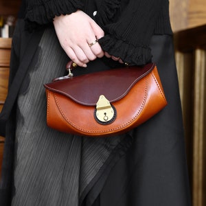 May include: A brown and burgundy leather handbag with a brass clasp and a curved shape. The bag has a short handle and is held by a person wearing a black sweater.