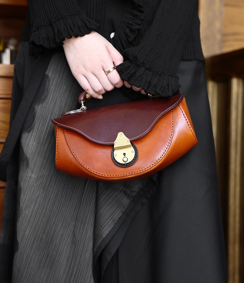 May include: A brown and burgundy leather handbag with a brass clasp and a curved shape. The bag has a short handle and is held by a person wearing a black sweater.