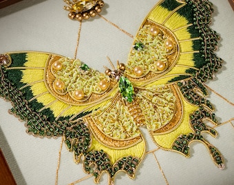Pode incluir: Um bordado emoldurado de uma borboleta com asas verdes e amarelas, decorado com contas e lantejoulas. A borboleta está centralizada na moldura, com uma pequena estrela dourada acima.