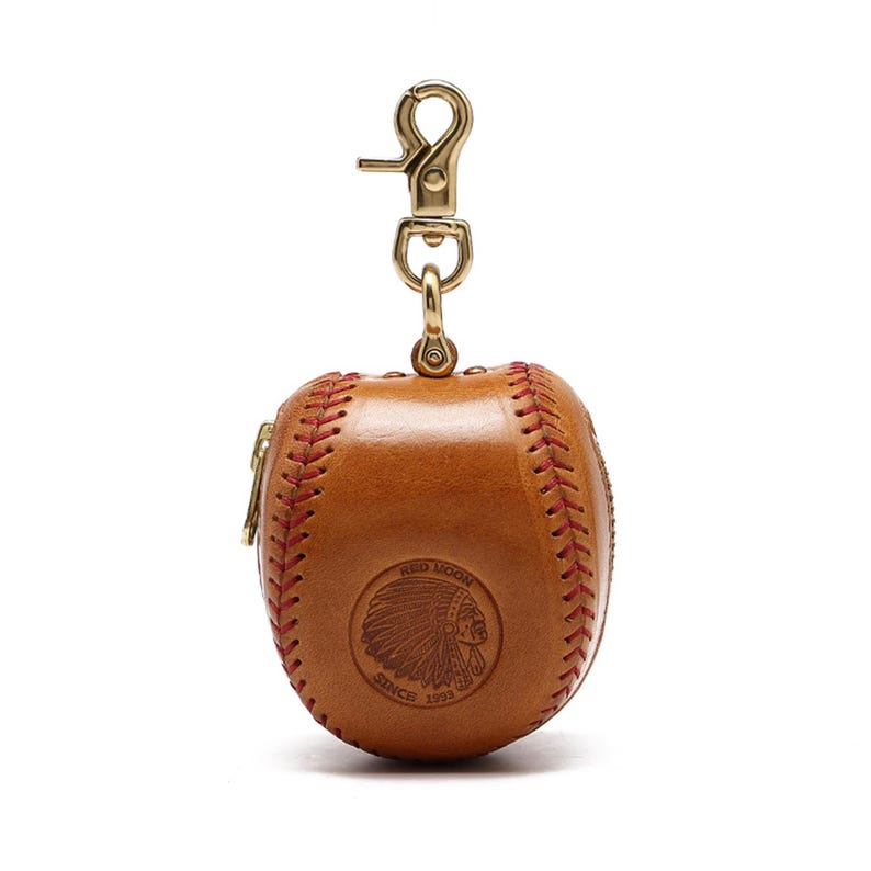 May include: A brown leather baseball-shaped coin purse with a red stitching detail and a gold zipper. The purse has a gold metal clasp and a logo that reads "Red Moon Since 1993" with an image of a Native American chief's head.