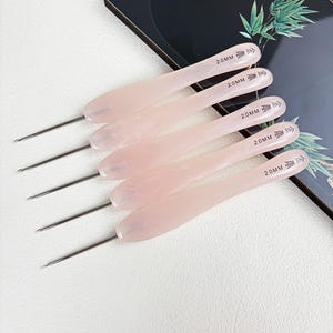Può includere: Un set di cinque uncinetti con manici in resina marmorizzata rosa chiaro. Ogni uncinetto presenta una punta in metallo argentato ed è chiaramente contrassegnato sul manico con la scritta 2.0MM e caratteri cinesi, indicando una misura uniforme.
