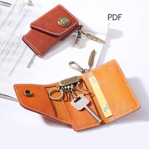 May include: A brown leather key holder with a key ring and a feather charm. The key holder has a flap that folds over to secure the keys. The key holder is open and shows a key, a credit card, and a key ring with multiple keys.