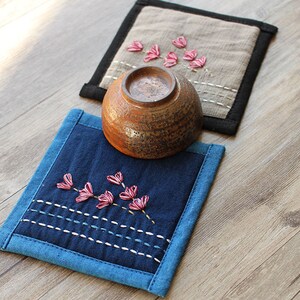 Puede incluir: Dos posavasos de tela hechos a mano con flores rosas bordadas. Los posavasos son de tela azul y beige con ribete negro. Un cuenco de cer&aacute;mica marr&oacute;n est&aacute; sentado encima de uno de los posavasos.