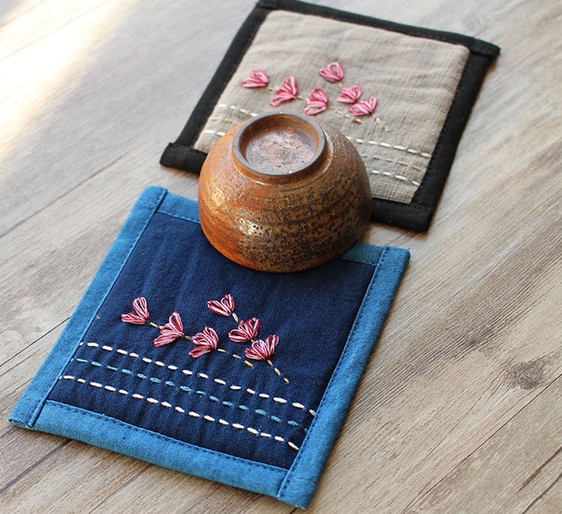 Puede incluir: Dos posavasos de tela hechos a mano con flores rosas bordadas. Los posavasos son de tela azul y beige con ribete negro. Un cuenco de cer&aacute;mica marr&oacute;n est&aacute; sentado encima de uno de los posavasos.