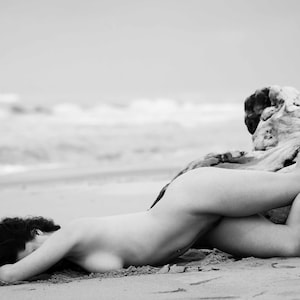 May include: A black and white photo of a person lying on a beach with their legs draped over a large piece of driftwood.