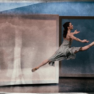May include: A ballerina wearing a light grey dress leaps through the air in a dance studio. The background is a faded blue and brown.