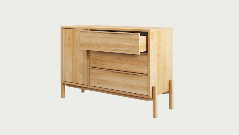 May include: A light wood dresser with three drawers and a cabinet door. The top drawer is open, revealing the wood interior. The dresser has four legs and a simple design.