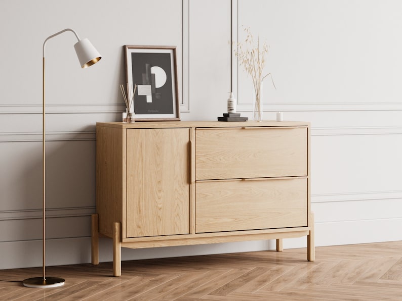 May include: A light wood cabinet with two drawers and a door. The cabinet has brass handles and sits on four legs. A framed black and white print with geometric shapes hangs above the cabinet.