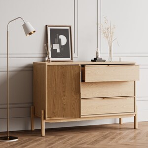 May include: A light wood dresser with three drawers and a cabinet door. The dresser has a gold metal floor lamp next to it. The dresser is decorated with a framed black and white print, a vase of dried flowers, and a bottle of lotion.