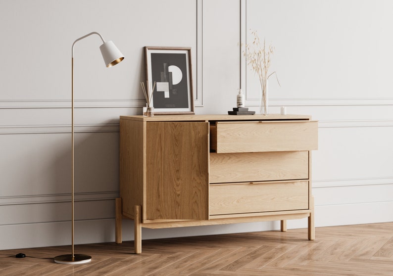 May include: A light wood dresser with three drawers and a cabinet door. The dresser has a gold metal floor lamp next to it. The dresser is decorated with a framed black and white print, a vase of dried flowers, and a bottle of lotion.