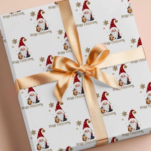 May include: A rectangular gift box wrapped in white paper featuring a repeating pattern of gnomes wearing red hats and holding mugs, interspersed with gold snowflakes and the text Merry Christmas, tied with a wide, shiny gold satin ribbon.