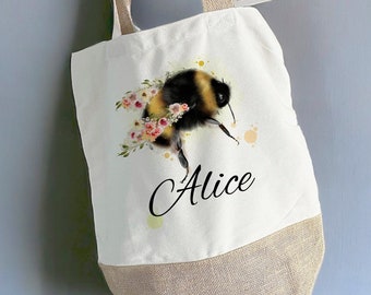 May include: A white canvas tote bag with a brown jute bottom. The bag features a watercolor illustration of a bumblebee with pink flowers and the name "Alice" printed in black cursive.