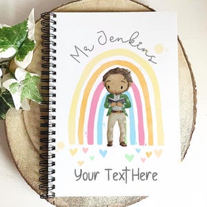 May include: Spiral-bound notebook with a colorful illustration of a teacher standing in front of a rainbow. The notebook has the text "Mr Jenkins" and "Your Text Here" on the cover. The notebook is resting on a wooden surface.