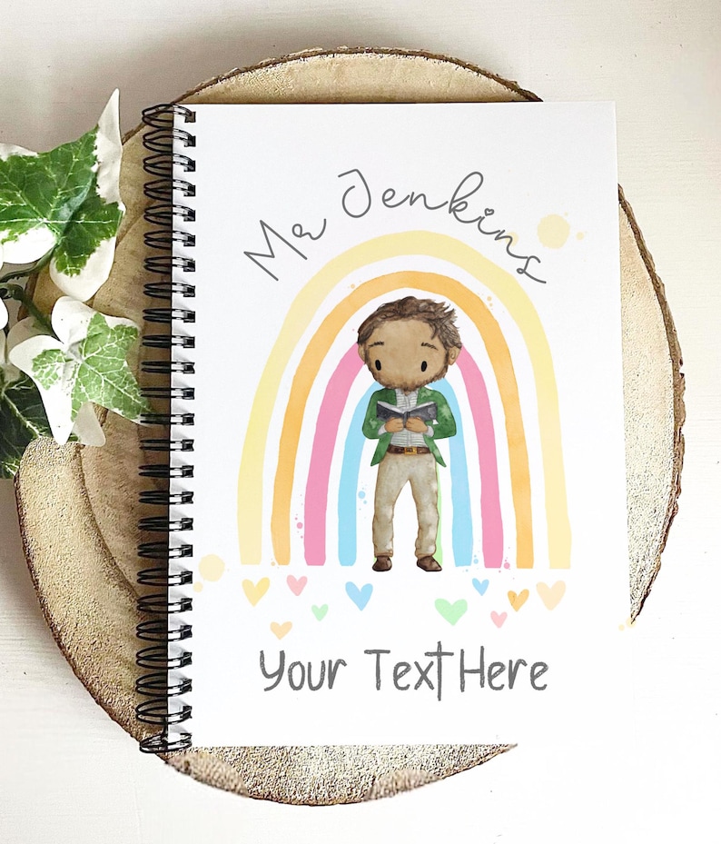 May include: Spiral-bound notebook with a colorful illustration of a teacher standing in front of a rainbow. The notebook has the text "Mr Jenkins" and "Your Text Here" on the cover. The notebook is resting on a wooden surface.