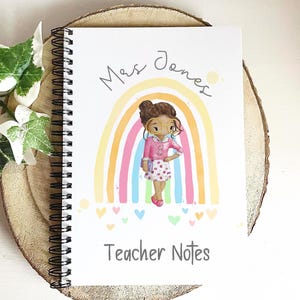 May include: A spiral-bound notebook with a colorful illustration of a teacher in front of a rainbow. The notebook cover has the text "Mrs. Jones" and "Teacher Notes" in a handwritten style. The notebook is on a wooden slice.