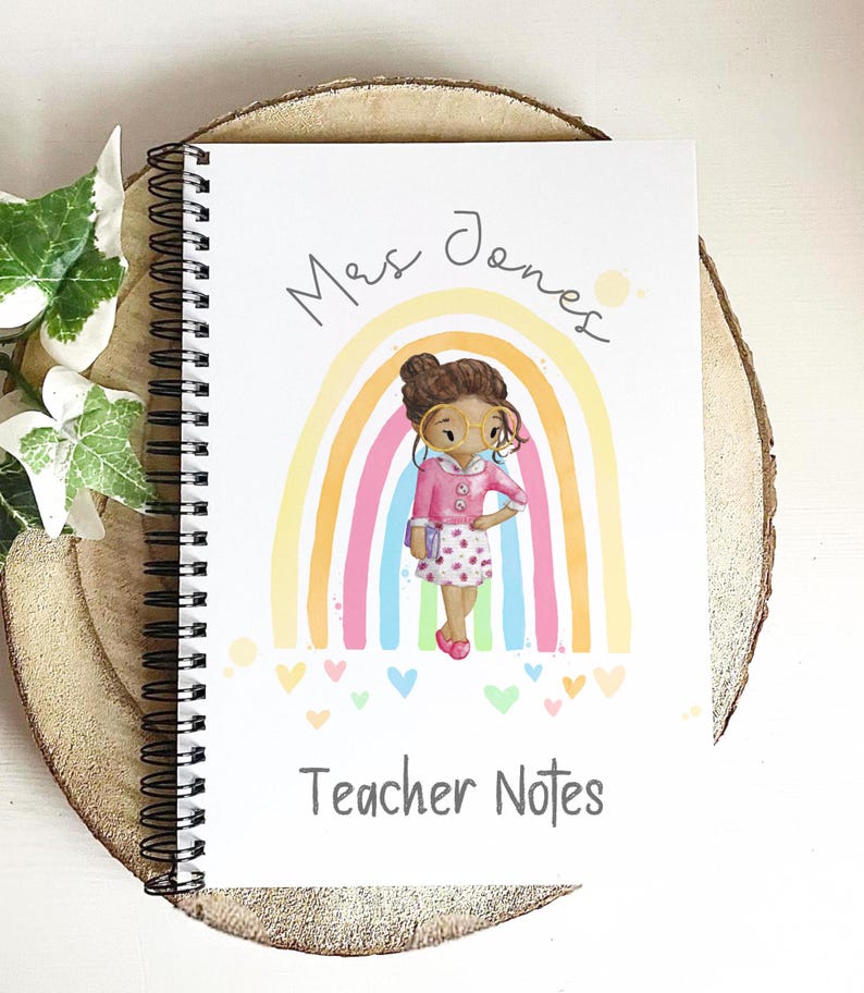 May include: A spiral-bound notebook with a colorful illustration of a teacher in front of a rainbow. The notebook cover has the text "Mrs. Jones" and "Teacher Notes" in a handwritten style. The notebook is on a wooden slice.