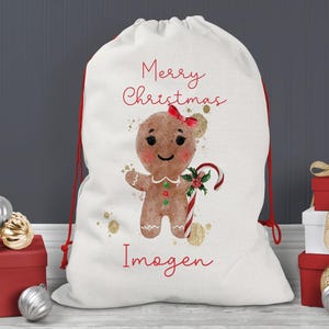 May include: A white Christmas gift bag with a red drawstring closure. The bag features a gingerbread person illustration with a red bow, candy cane, and the text "Merry Christmas" and a name in red script. Gold accents are scattered around the design.