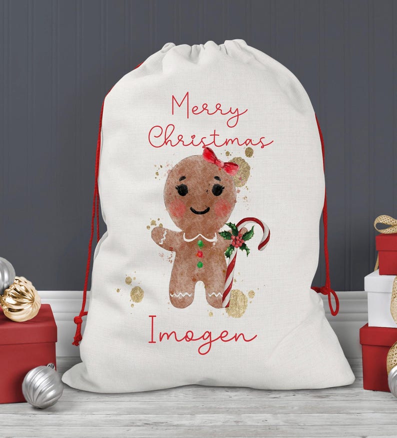 May include: A white Christmas gift bag with a red drawstring closure. The bag features a gingerbread person illustration with a red bow, candy cane, and the text "Merry Christmas" and a name in red script. Gold accents are scattered around the design.