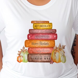 May include: A white t-shirt featuring a watercolour design of a stack of books with autumn-themed words on the spines. The words are "Cozy Jumpers", "Hot Chocolate", "Toasted Marshmallows", "Halloween", "Warm Blankets", "Crispy Leaves", and "Pumpkin Spice". There are also two pumpkins and leaves at the bottom of the design.