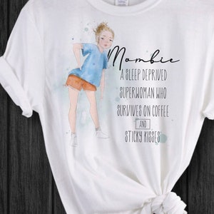 Peut inclure: T-shirt blanc avec une illustration &agrave; l'aquarelle d'une personne en chemise bleue et short orange. Le t-shirt porte le mot "Mombie" et le texte "Une superwoman priv&eacute;e de sommeil qui survit gr&acirc;ce au caf&eacute; et aux bisous collants."