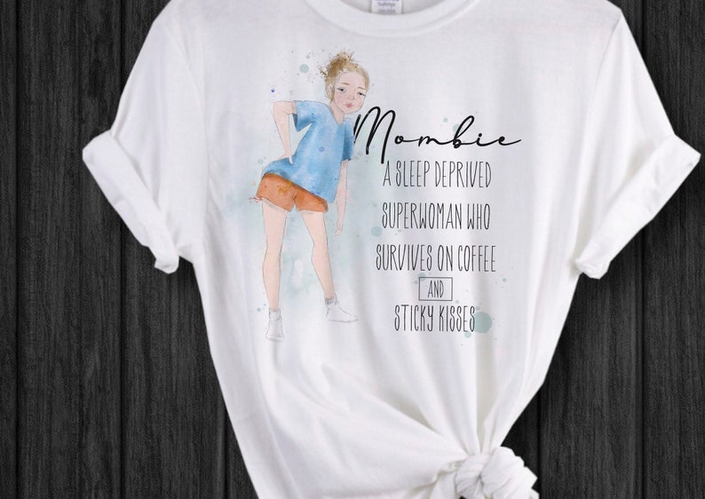 Peut inclure: T-shirt blanc avec une illustration &agrave; l'aquarelle d'une personne en chemise bleue et short orange. Le t-shirt porte le mot "Mombie" et le texte "Une superwoman priv&eacute;e de sommeil qui survit gr&acirc;ce au caf&eacute; et aux bisous collants."