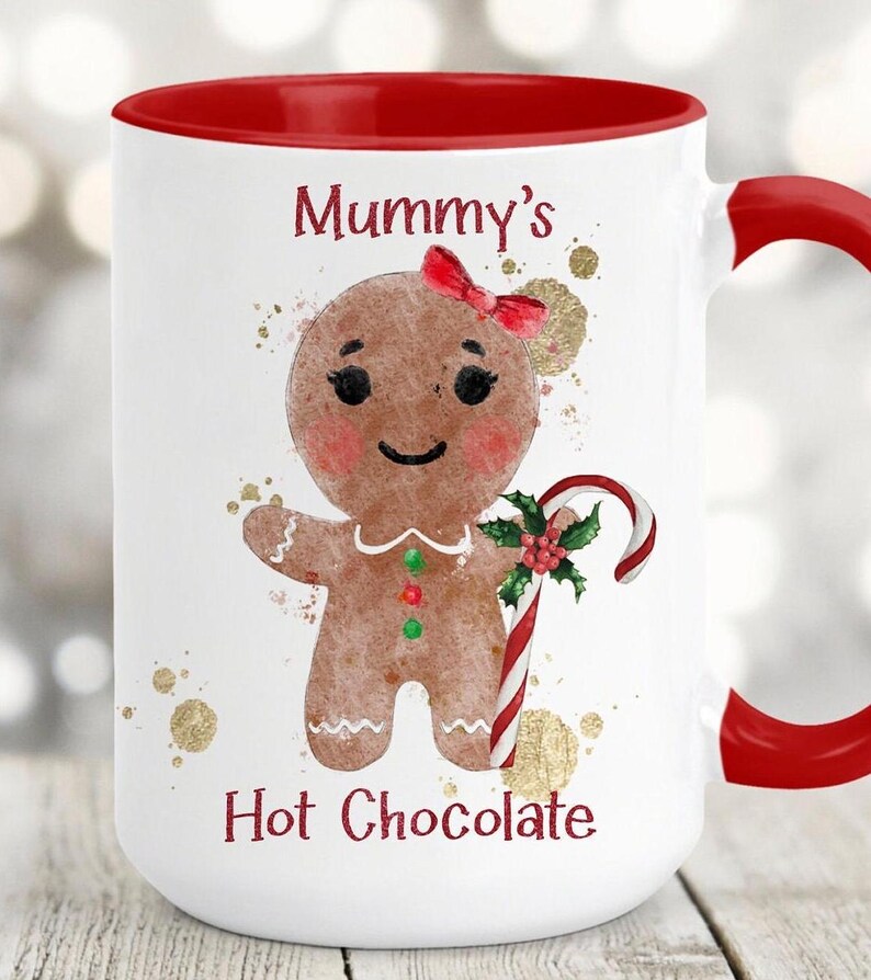 May include: White ceramic mug with a red interior and handle. The mug features a gingerbread person illustration with a red bow, holding a candy cane. The text "Mummy's Hot Chocolate" is printed on the mug.