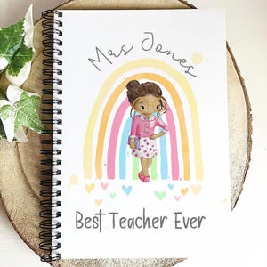 May include: Spiral-bound notebook with a colorful illustration of a teacher in front of a rainbow. The notebook cover has the text "Mrs Jones" and "Best Teacher Ever" in a handwritten font. The notebook is on a wooden surface.