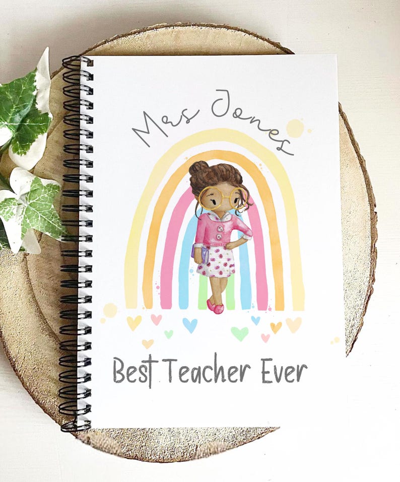 May include: Spiral-bound notebook with a colorful illustration of a teacher in front of a rainbow. The notebook cover has the text "Mrs Jones" and "Best Teacher Ever" in a handwritten font. The notebook is on a wooden surface.