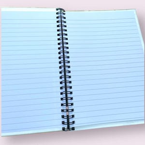 May include: An open notebook with lined, light blue pages. The pages are held together by a black metal spiral binding. The notebook is open, revealing the blank pages ready for writing or sketching. The notebook's cover is a light beige color.