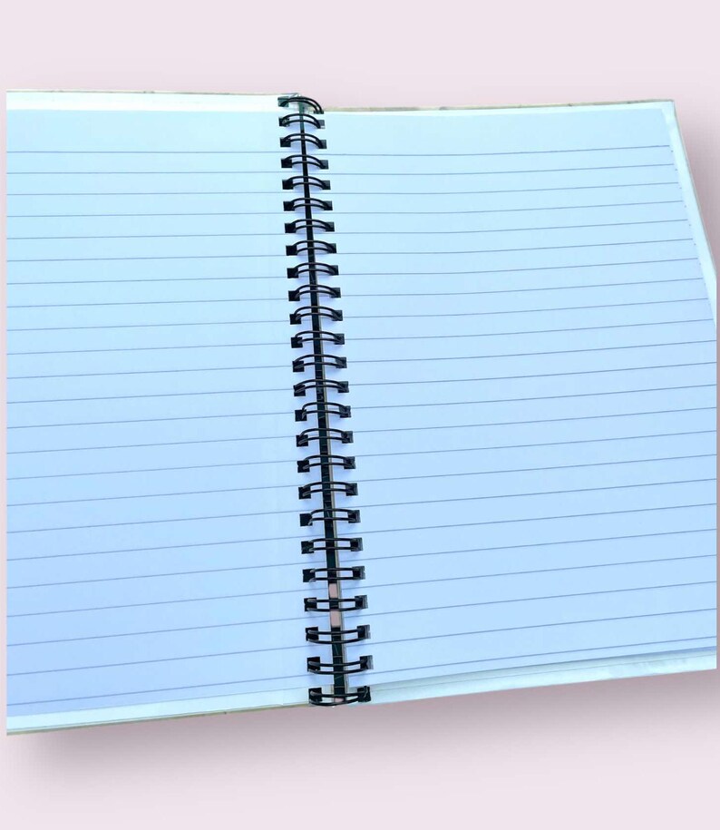 May include: An open notebook with lined, light blue pages. The pages are held together by a black metal spiral binding. The notebook is open, revealing the blank pages ready for writing or sketching. The notebook's cover is a light beige color.