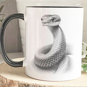 May include: White ceramic mug with a black handle and rim. The mug showcases a detailed grayscale illustration of a snake in a coiled pose. The snake's scales and features are meticulously rendered, creating a realistic effect.