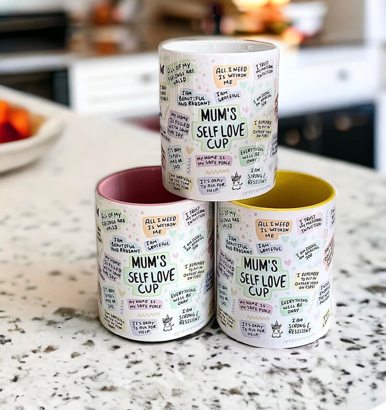May include: Three ceramic mugs with a white background and colorful text. The mugs have a pink, white, and yellow rim. The text on the mugs reads "Mum's Self Love Cup" and includes affirmations such as "All of my feelings are valid", "I am beautiful and radiant", "My home is filled with love", "It's okay to ask for help", "I trust my intuition", "I remember to put my oxygen mask on first", "Everything will be okay", "I am strong and resilient".