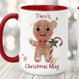 May include: White ceramic Christmas mug with a red handle and interior. The mug features a gingerbread man illustration with a candy cane and the text "Theo's Christmas Mug". Gold paint splatters are in the background.