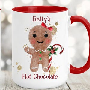 May include: White ceramic mug with a red interior and handle. The mug features a gingerbread person with a red bow, holding a candy cane. The text "Betty's Hot Chocolate" is printed on the mug, along with gold accents.