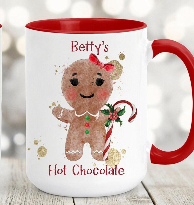 May include: White ceramic mug with a red interior and handle. The mug features a gingerbread person with a red bow, holding a candy cane. The text "Betty's Hot Chocolate" is printed on the mug, along with gold accents.
