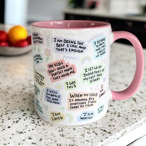 May include: White ceramic mug with a pink rim and handle. The mug is covered in colorful, handwritten affirmations, including "I am doing the best I can, and that's enough", "My kids don't need a perfect mum: they need me", "I got this", "I am loved", "I am worthy", and "When my child is having big emotions I will share my calm".