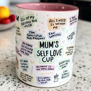 May include: White ceramic mug with a pink rim, featuring colorful text and illustrations. The text includes phrases like "All of my feelings are valid", "Mum's Self Love Cup", and "It's okay to ask for help".