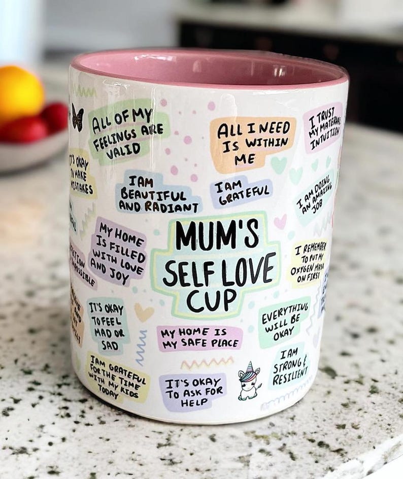 May include: White ceramic mug with a pink rim, featuring colorful text and illustrations. The text includes phrases like "All of my feelings are valid", "Mum's Self Love Cup", and "It's okay to ask for help".