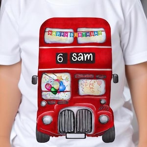 May include: A white t-shirt featuring a red double-decker bus graphic. The bus has a banner that reads "Happy Birthday" and a sign that says "6 Sam".
