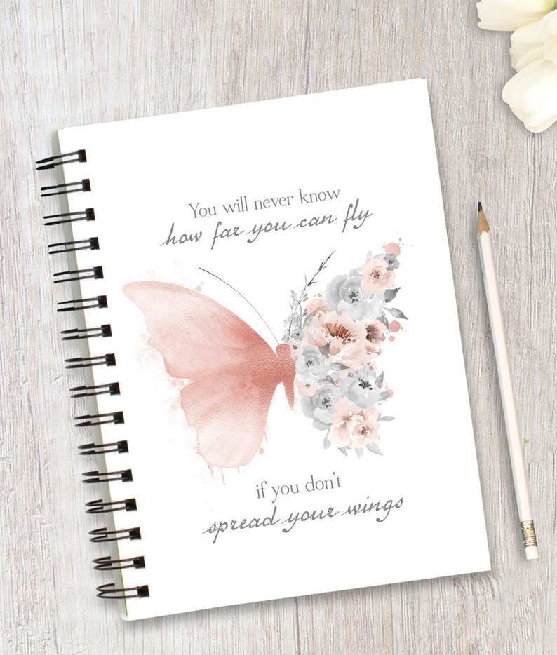 May include: A white notebook with a spiral binding features a watercolor illustration of a pink butterfly with gray and pink flowers. The notebook has the text "You will never know how far you can fly if you don't spread your wings".