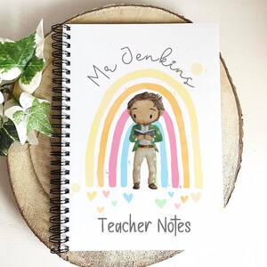 May include: Spiral-bound notebook with a colorful rainbow design and the text "Mr Jenkins" and "Teacher Notes." The cover features an illustration of a teacher reading a book in front of a rainbow. The notebook is resting on a wooden surface.