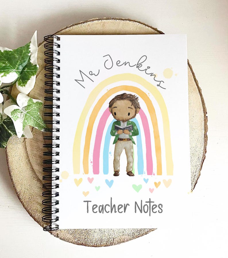 May include: Spiral-bound notebook with a colorful rainbow design and the text "Mr Jenkins" and "Teacher Notes." The cover features an illustration of a teacher reading a book in front of a rainbow. The notebook is resting on a wooden surface.