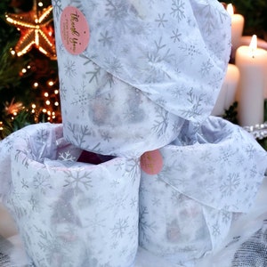 May include: Three cylindrical containers wrapped in white paper with a silver snowflake pattern. Each container is sealed with a pink sticker that reads "Thank you for your order". The background includes a Christmas tree and lit candles.