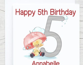 以下が含まれることがあります： 「Happy 5th Birthday」の赤い文字が書かれた白いバースデーカード。赤い傘とブーツを履いた黄色いアヒルのイラストが描かれています。「Annabelle」という名前も赤色で書かれています。