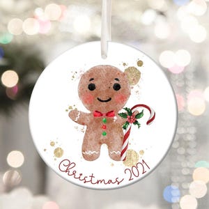 May include: A round Christmas ornament featuring a gingerbread man with a red bow tie and candy cane. The ornament has the text "Christmas 2021" in red script, with gold accents. A white ribbon is attached for hanging.