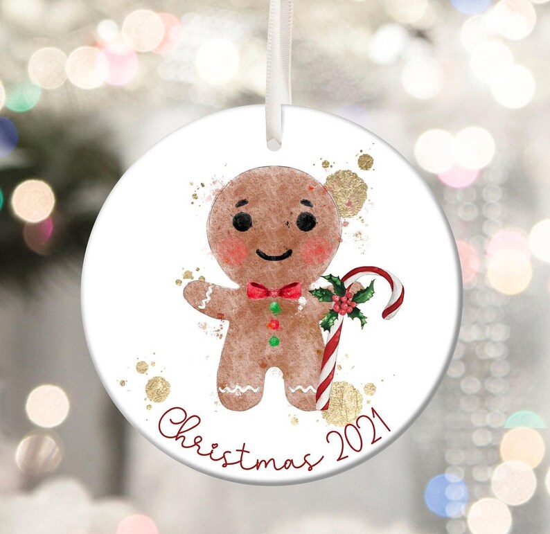 May include: A round Christmas ornament featuring a gingerbread man with a red bow tie and candy cane. The ornament has the text "Christmas 2021" in red script, with gold accents. A white ribbon is attached for hanging.