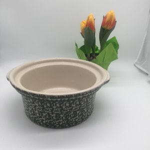 May include: A ceramic slow cooker with a cream-coloured interior and a dark green speckled exterior. The rim is cream-coloured. The slow cooker is on a white surface, with a small pot of yellow and orange tulips in the background.