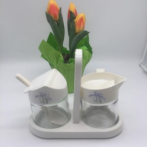 Puede incluir: Un juego de condimentos blanco con asa, con dos tarros de vidrio con tapas y cucharas blancas, y una jarrita. Cada pieza tiene un diseño floral morado. Un pequeño tiesto de tulipanes naranjas y amarillos está al fondo.